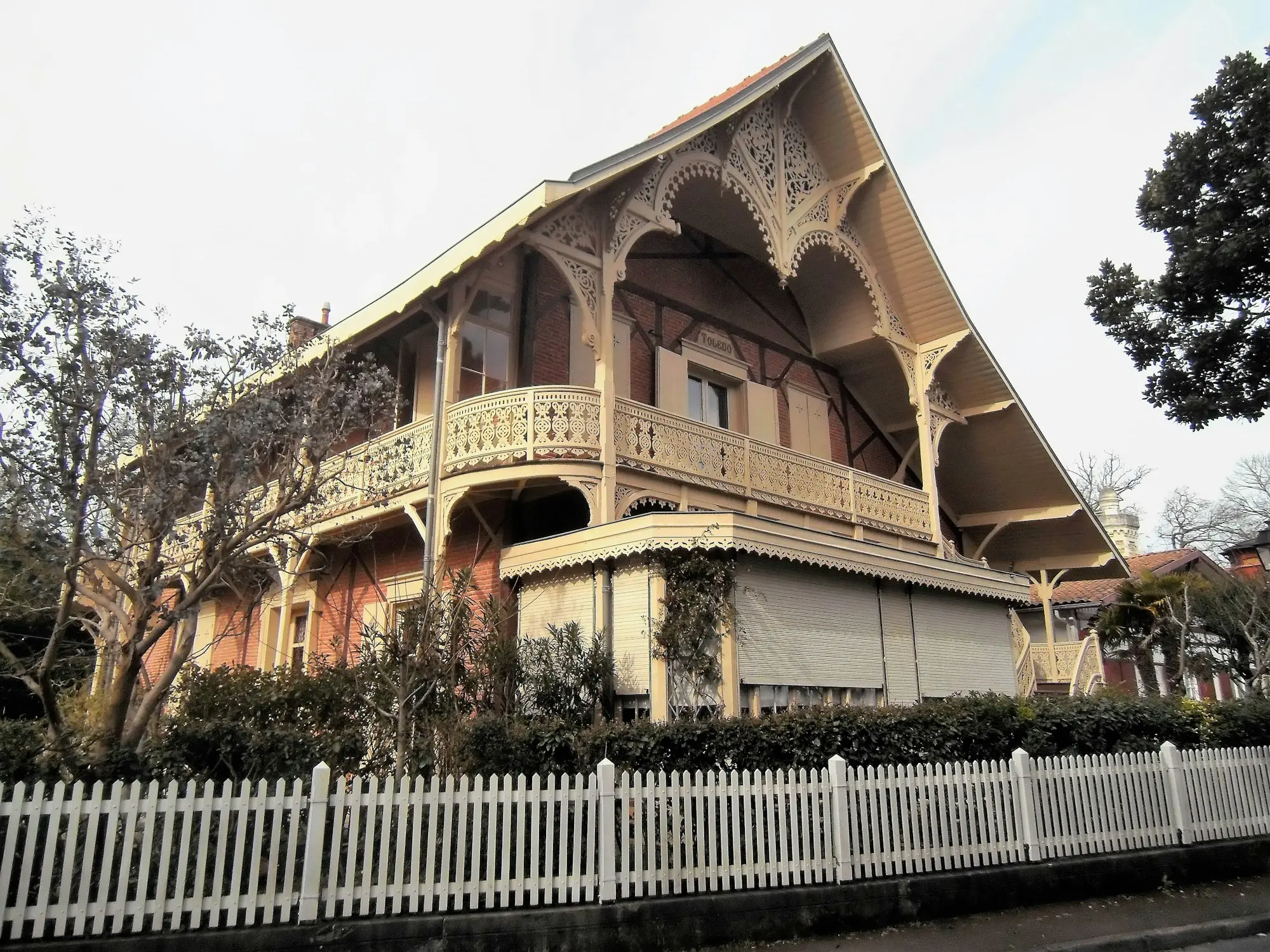 Brick house with ornate timber trim and a balcony behind a white fence