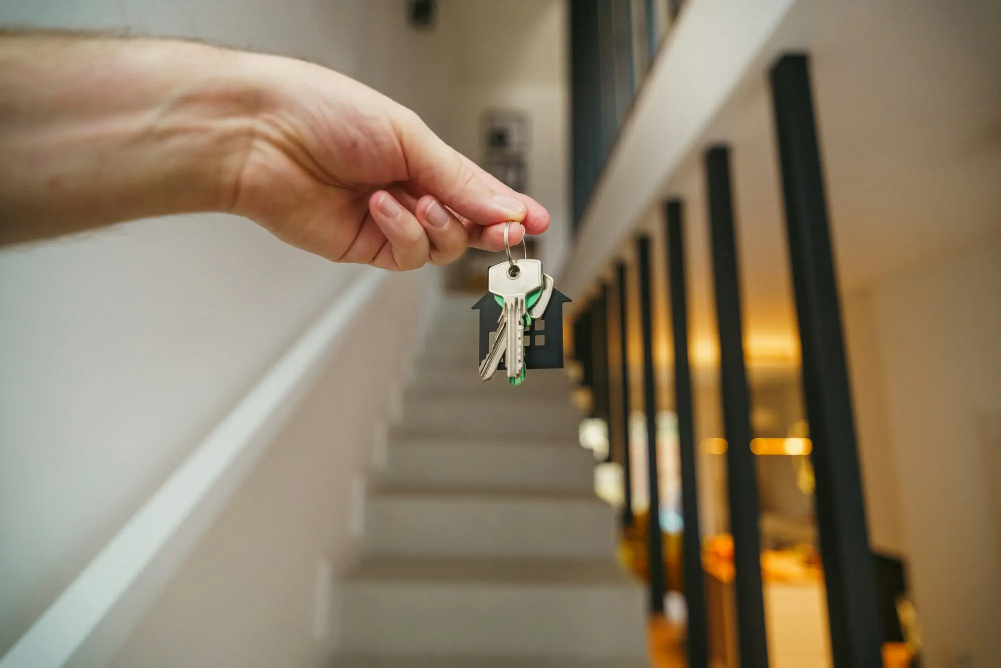 Hand holding keys with a house-shaped keyring in front of a staircase